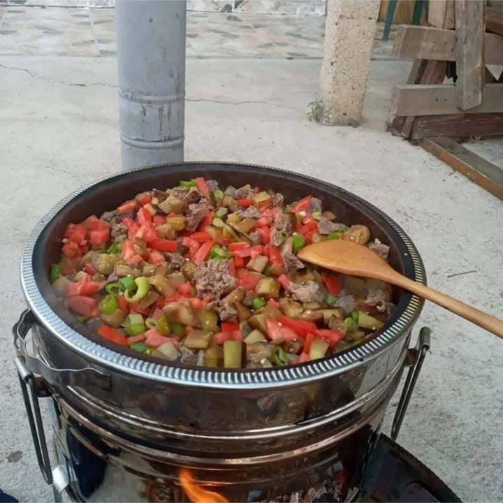 A HAKAN Portable Guvec Mangal Soba Clay Grill Barbecue Stove 15-inch for outdoor camping is filled with chopped vegetables and meat, including tomatoes and green peppers, while a wooden spoon rests on its rim. It sits outdoors on concrete, framed by wood and metal in the background.