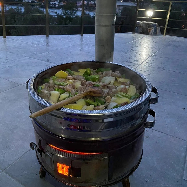 A large HAKAN Portable Guvec Mangal Soba Clay Grill Barbecue Stove 15-inch for outdoor camping sits filled with chopped vegetables and meat, steaming as a wooden spoon rests on its edge. The background reveals a railing and distant lights under the evening sky.