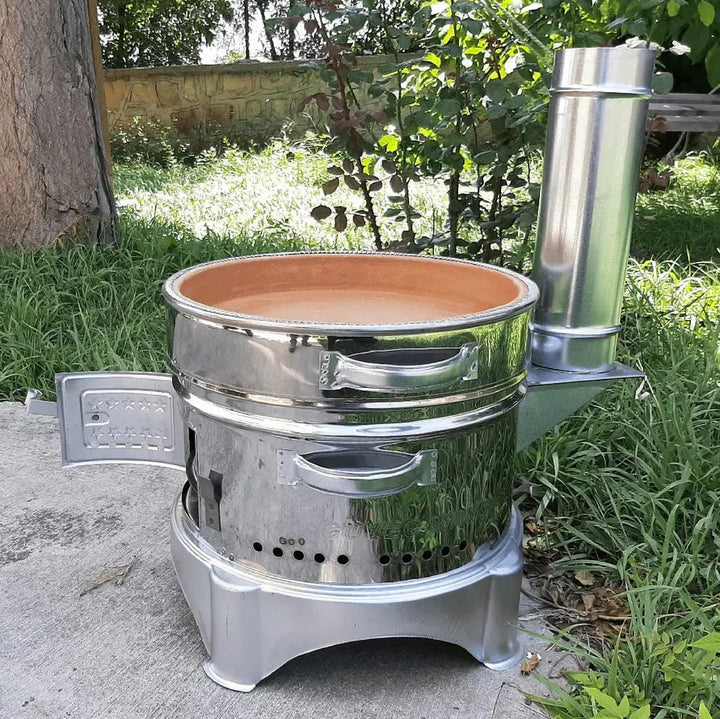 A HAKAN Portable Guvec Mangal Soba Clay Grill Barbecue Stove, 15-inch, stands outdoors with a clay casserole pot on its shiny metal surface; it includes a side door, chimney pipe, and handles, set against tree and foliage like a Turkish barbecue.