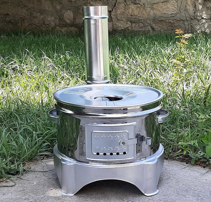 A HAKAN Portable Guvec Mangal Soba Clay Grill Barbecue Stove, 15-inch for Outdoor Camping, sits on a concrete surface. The shiny chrome stove features a round body, small chimney, and front door with air vents. The scene includes a grass lawn and stone wall in the background.