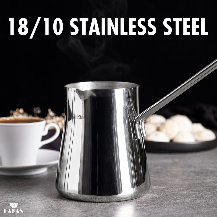 A polished 18/10 stainless steel Turkish coffee pot with a long handle on a countertop, with a coffee cup and sweets in the background