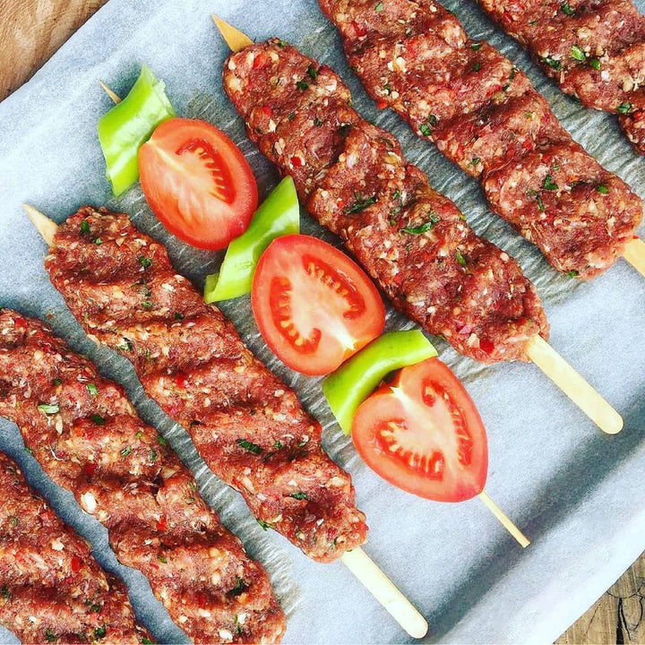 Close-up of six raw minced meat kebabs on Hakan 9.8-inch BBQ-grade wooden skewers, placed on parchment paper. Two skewers are garnished with sliced green bell peppers and halved tomatoes for a pop of color.