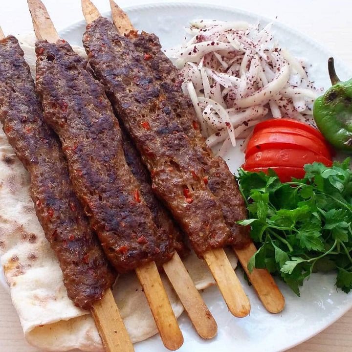 On a plate, four HAKAN skewers hold BBQ delights, accompanied by warm flatbread, sliced onions, juicy tomato slices, crisp green pepper, and fresh cilantro.