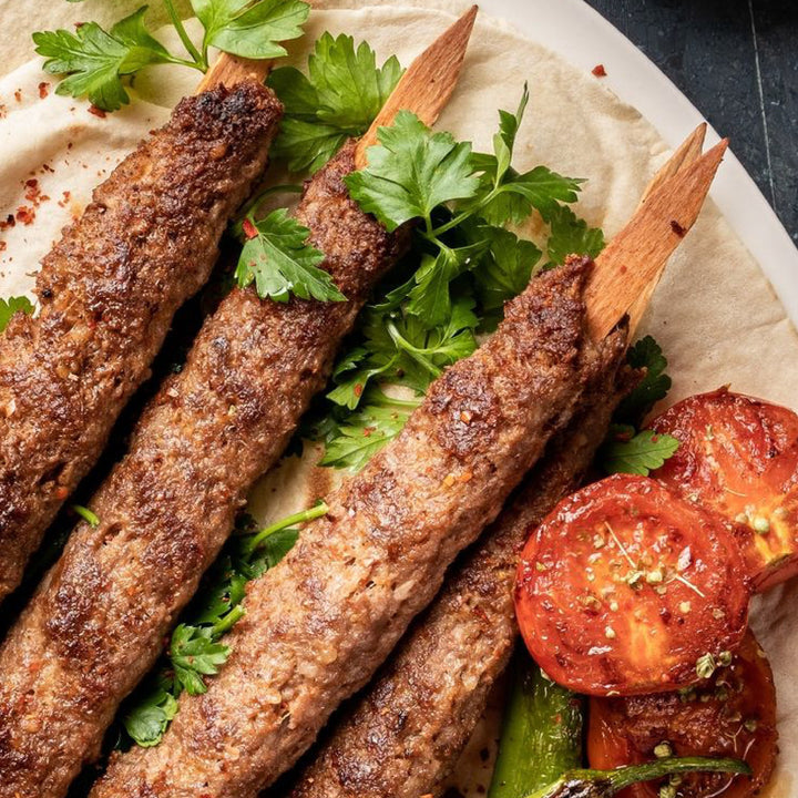 Three grilled kebabs on HAKAN 9.8-inch wooden flat skewers rest on flatbread, garnished with parsley. Seasoned tomatoes and green peppers accompany the dish, presented elegantly on a round white plate.